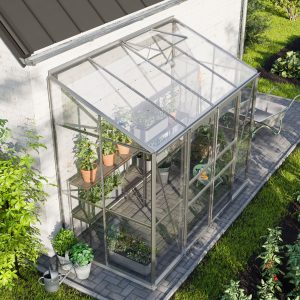 Bright aluminum greenhouse with glass panels on a garden patio, surrounded by lush plants and soil beds, ideal for versatile gardening and plant cultivation.