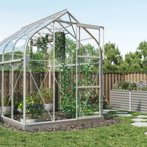 Aluminum greenhouse with glass panels in a landscaped garden, featuring vegetable plants, flowers, and a wooden fence for privacy, ideal for home gardening and plant cultivation.