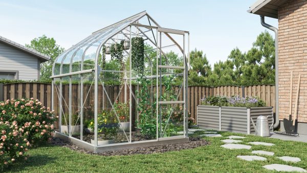 Aluminum greenhouse with glass panels in a landscaped garden, featuring vegetable plants, flowers, and a wooden fence for privacy, ideal for home gardening and plant cultivation.